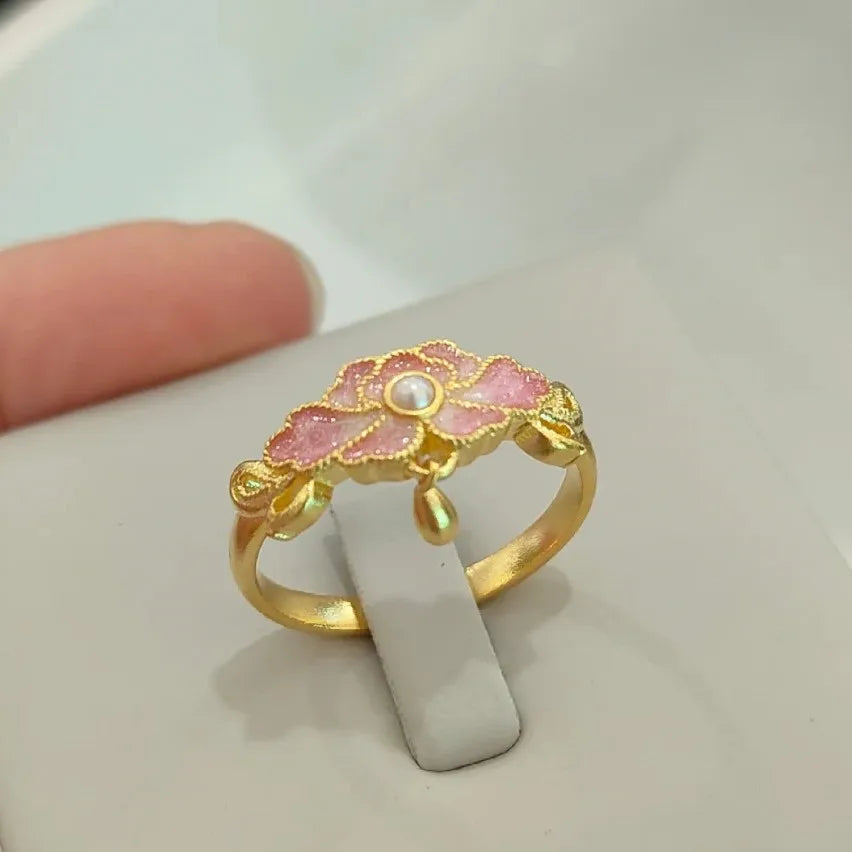 Gold ring with pink floral design and pearl center on a white stand.